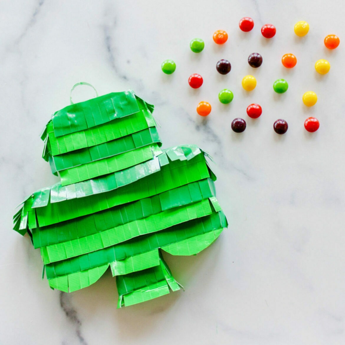 A white marble background with a shamrock covered with duct tape fringe next to multi colored candy